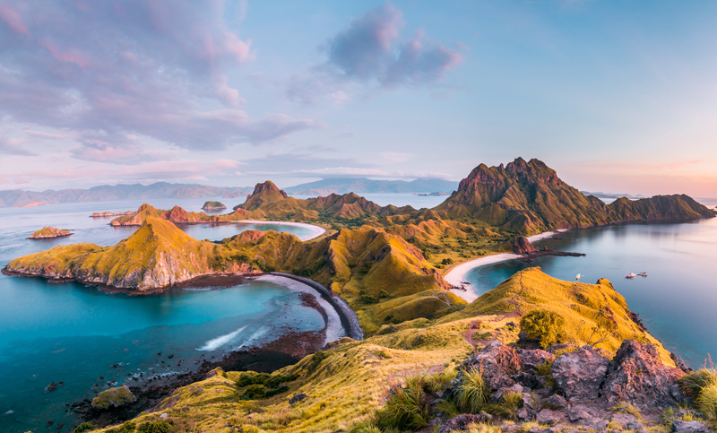 Rolling, green hills form a rugged coastline, surrounded by turquoise waters with small islands dotted in the distance, under a sky painted with soft clouds and warm sunlight.