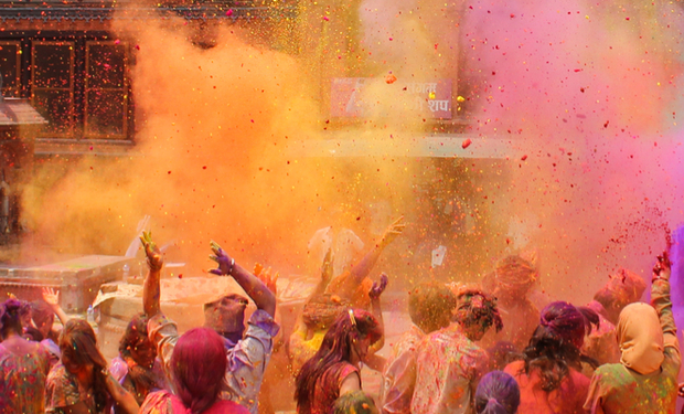 People joyfully throw vibrant colored powders into the air during a festive celebration, surrounded by buildings and a partially visible vehicle. Signs in the background feature text in a regional script.