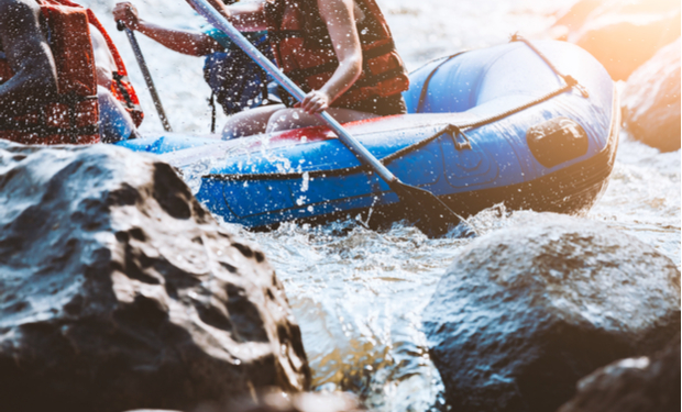 A blue inflatable raft navigates through rocky, splashing rapids. People inside, wearing red life jackets, paddling with oars amid sunlight filtering through the scene.
