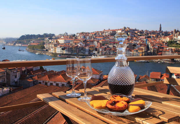 Two wine glasses, wine and Portuguese pastel de nata on a table outside, looking over a port in Portugal on a sunny day.