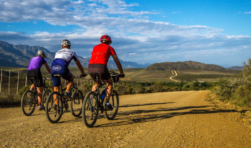 Three cyclists ride on a gravel road wearing colorful jerseys, surrounded by open landscape and distant mountains under a partly cloudy sky.