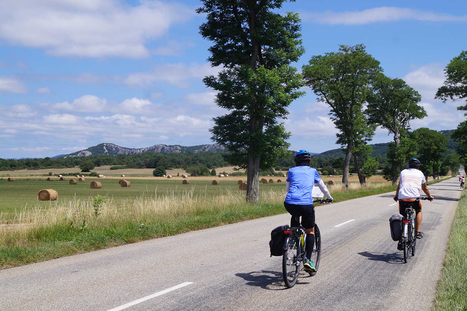 Cyclists ride on a rural road flanked by hay bales and trees, mountains visible in the distance under a bright, partly cloudy sky.