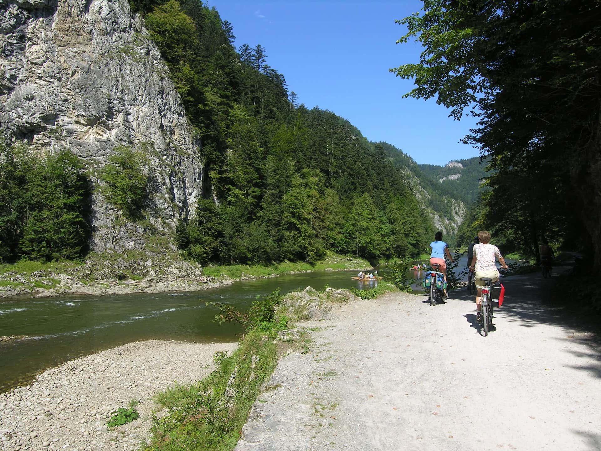 Two people ride bicycles along a riverside path in a forested mountain area, with rocky cliffs and a blue sky. In the river, there are people rafting downstream.