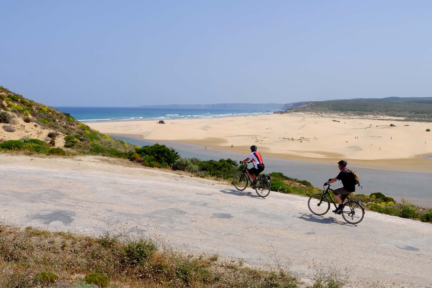 Two cyclists ride along a paved road. The backdrop features a sandy beach, blue ocean, and distant hills under a clear sky. Vegetation lines the road.