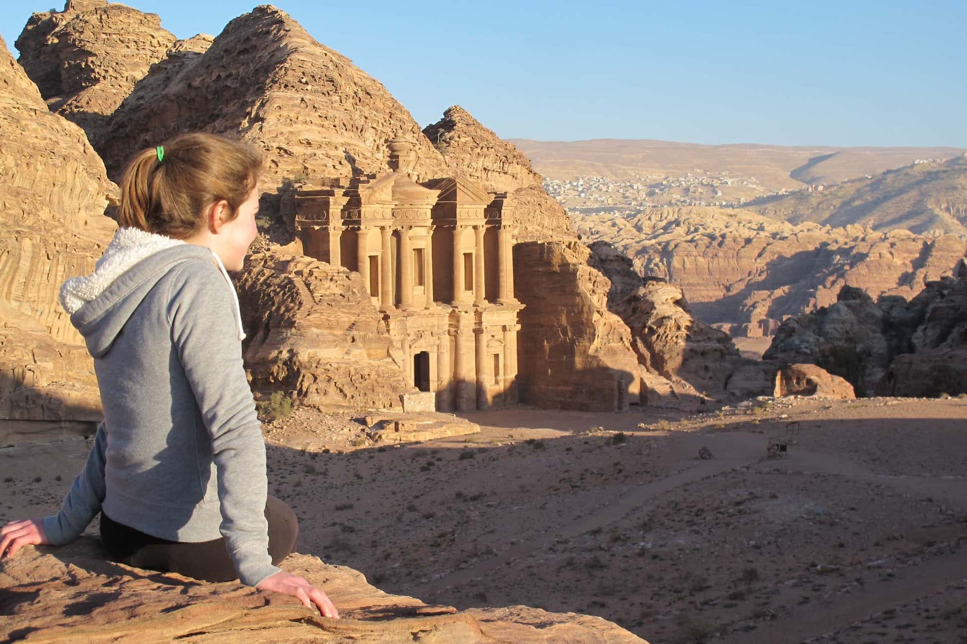 A person sits on a rock ledge overlooking Petra's ancient stone façade carved into a rugged, desert landscape under a clear blue sky.