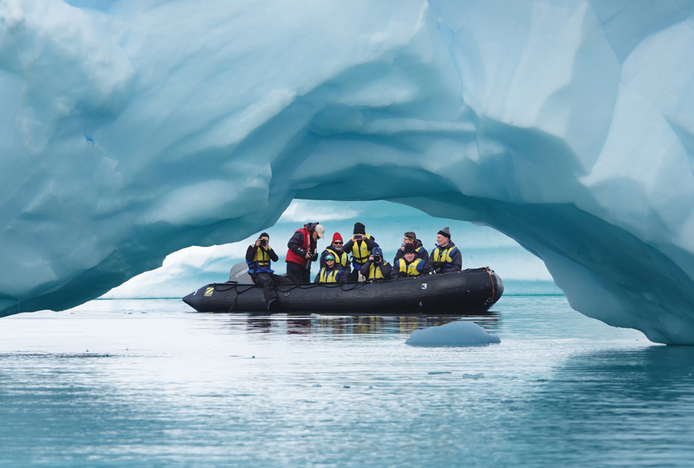 A black inflatable raft carries people in yellow life jackets calmly passing under an arching blue iceberg in icy, serene waters.