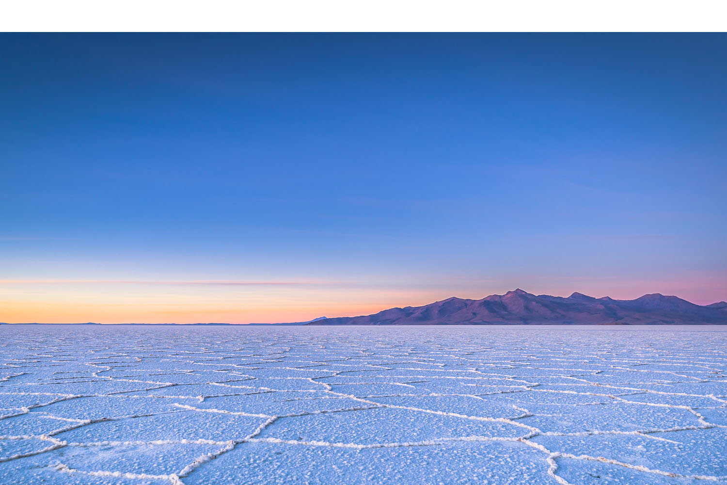 Cracked salt flats stretch across the foreground, meeting distant mountains under a clear blue and orange-gradient sky at sunset, creating a serene and expansive landscape.