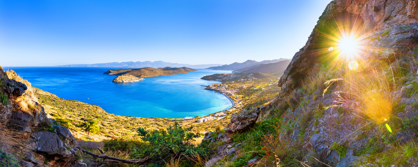 Sun shining through a rocky cliff, illuminating a vibrant coastal landscape with a vivid blue sea and distant mountains under a clear sky.