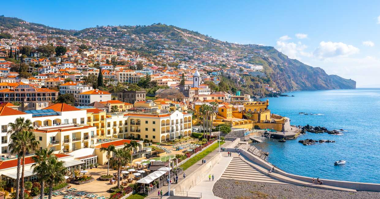 Colorful buildings with red-tiled roofs cascade down a lush hillside towards a calm, blue ocean. A few people stroll along the waterfront promenade beneath a bright, clear sky.