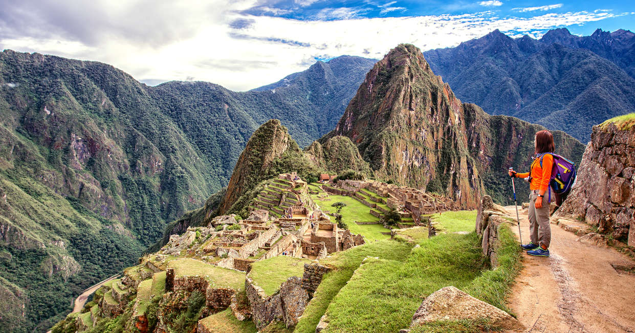 A person stands holding a hiking pole, overlooking the ancient Incan ruins of Machu Picchu, surrounded by lush green mountains under a partly cloudy sky.