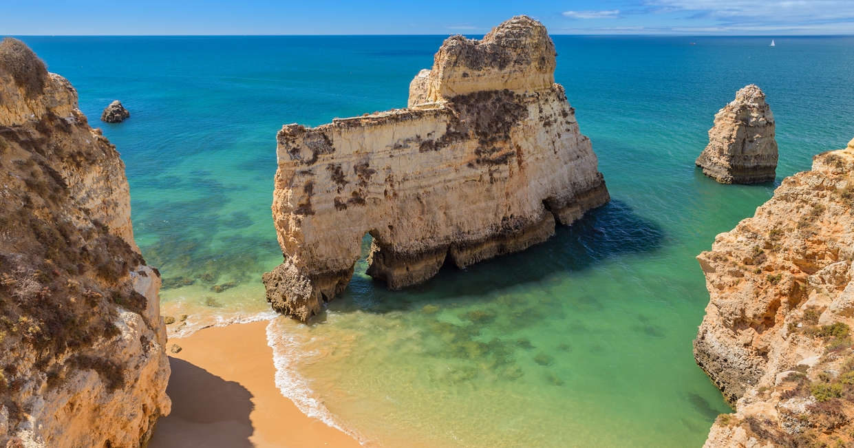 Rock formations rise dramatically from clear turquoise water, surrounded by sandy beaches and more rock cliffs under a bright blue sky, depicting a serene coastal scene.