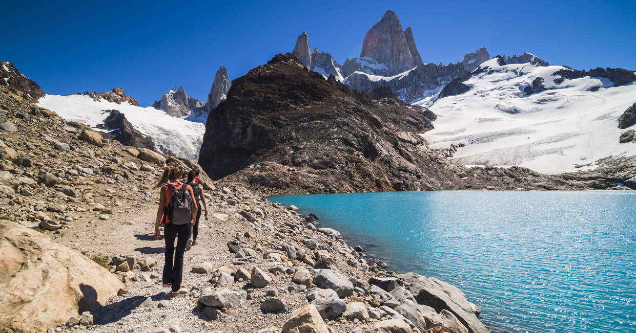 Hikers walk on a rocky trail beside a turquoise mountain lake, surrounded by snowy peaks under a clear blue sky.