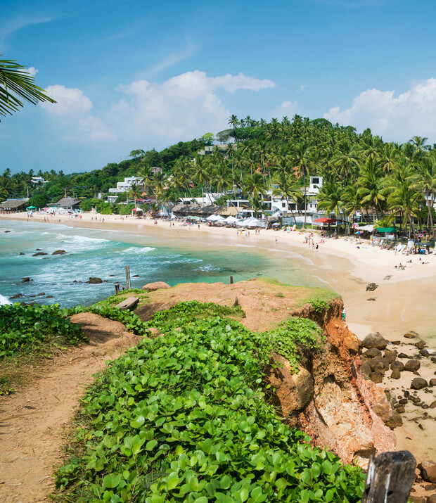 A sandy beach with people enjoying the sun, coconut trees, and surrounding lush greenery; white buildings dot the hillside under a clear blue sky with scattered clouds.