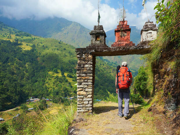 A hiker carrying a large orange backpack walks beneath a stone arch adorned with small flags, overlooking a lush, mountainous valley under a partly cloudy sky.
