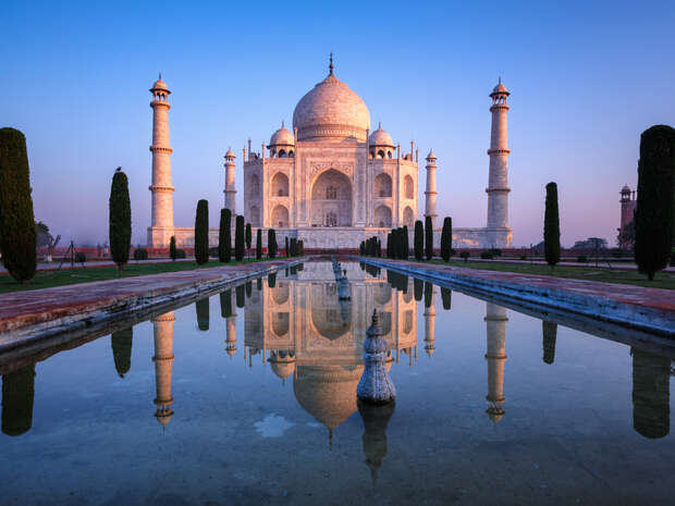 The Taj Mahal, a white marble mausoleum with large dome and four minarets, reflects in a still reflecting pool, surrounded by trees, during a clear, serene sunrise.