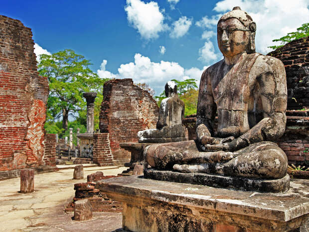 A weathered stone Buddha statue sits serenely on a pedestal amidst ancient, crumbling brick ruins, under a clear blue sky with scattered clouds and surrounding green foliage.