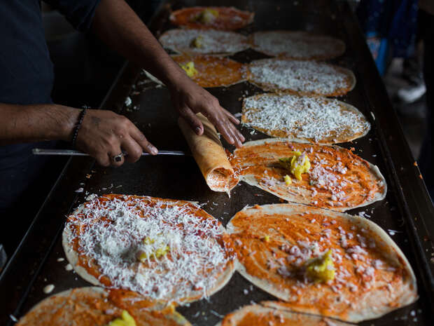 A person rolls a dosai filled with cheese and potato filling on a large griddle, surrounded by multiple flat, open dosais topped with similar ingredients in a bustling kitchen setting.