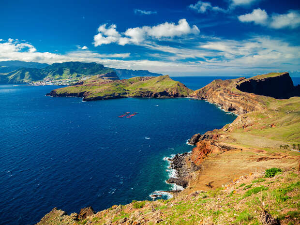 Coastal cliffs stretch into the ocean, with rugged terrain and green hills meeting deep blue water. Bright skies dotted with clouds add a vibrant backdrop to the scenic landscape.