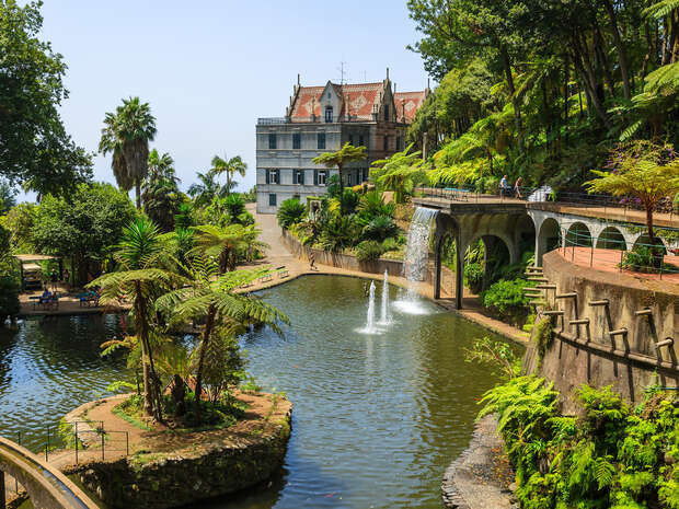 A historic manor overlooks a lush garden with a pond featuring fountains. Visitors walk along an arched bridge, surrounded by tropical plants and palm trees on a sunny day.