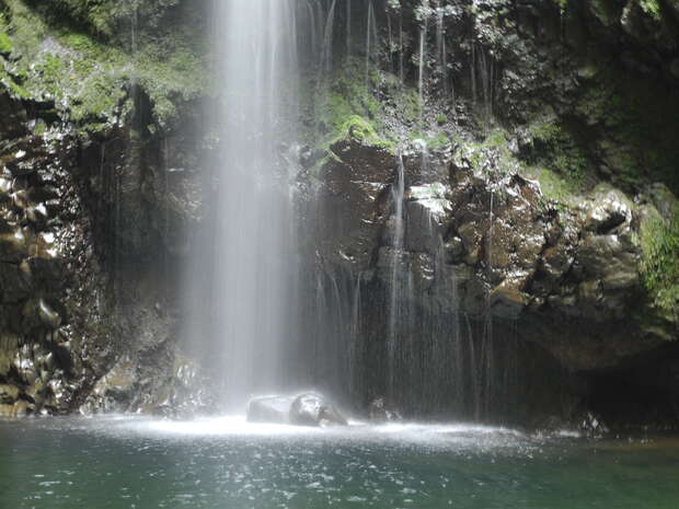 Waterfall cascades into a rocky pool, creating mist and ripples; surrounded by moss-covered rocks and lush green vegetation, conveying a serene, natural environment.