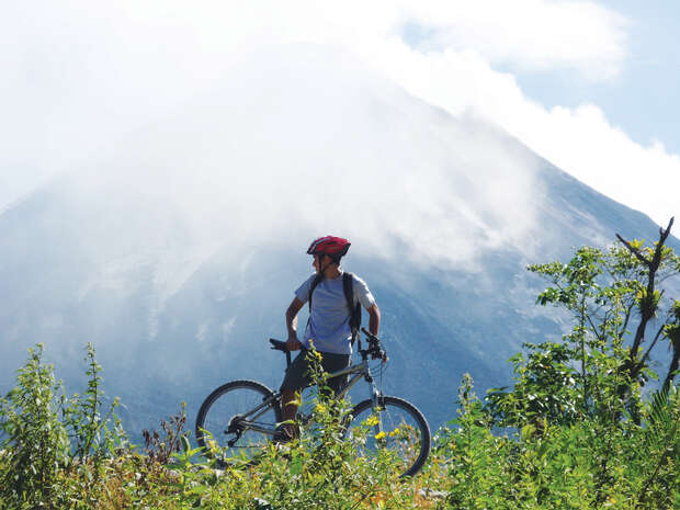 A person wearing a helmet is standing with a bicycle, surrounded by lush greenery, against the backdrop of a mist-covered mountain.