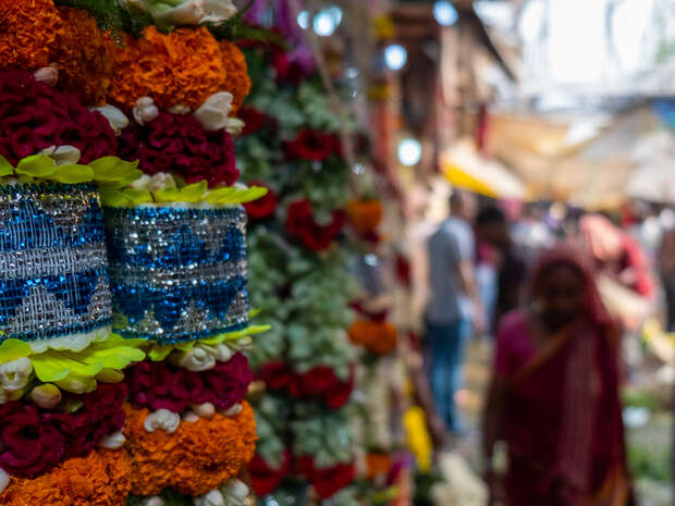 Garlands of vibrant orange, red, and white flowers hang in front, with sparkling blue decorations. A busy market scene is in the background, populated by blurred figures and market stalls.