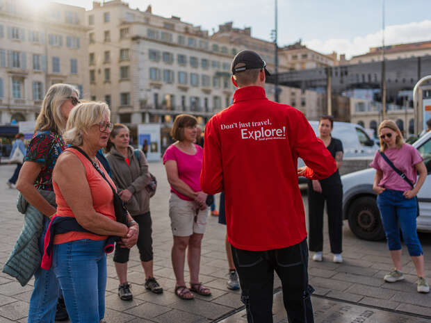 A tour guide, wearing a red jacket with the text "Don't just travel. Explore!", talks to a group of people in a sunny, urban setting with historical buildings.