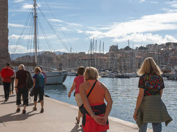 People walk along a waterfront path, with a sailing ship docked nearby and a marina filled with sailboats. In the background, a cityscape rises under a partly cloudy sky.