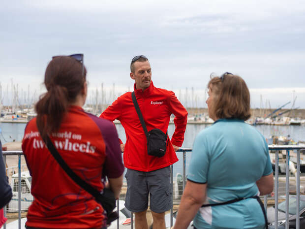 A man in a red jacket labeled "Explore" talks to three people outdoors. They stand by a marina with numerous sailboats and a cloudy sky in the background.