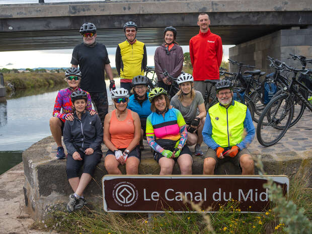 Cyclists sit and stand near bicycles, smiling under a bridge by a canal. They wear helmets and colorful cycling attire. A sign reads “Le Canal du Midi.”
