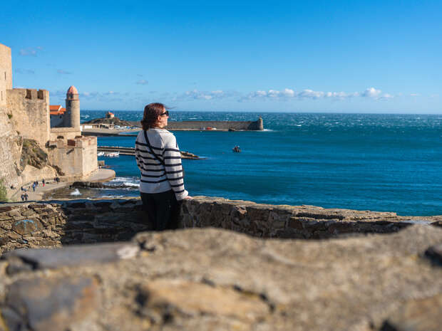 A person wearing a striped sweater stands on a stone wall, gazing at the ocean, with historic buildings and a lighthouse in the background under a clear blue sky.