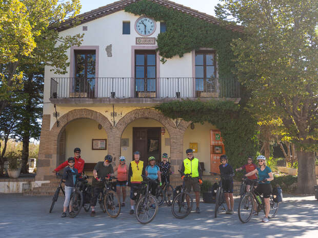 Cyclists stand with bikes in front of a two-story building with arched windows, a clock, and ivy. Trees frame the scene. A sign reads "RELOJ" above the entrance.