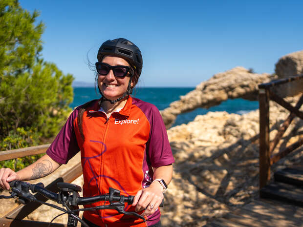 A cyclist stands smiling with a mountain bike, wearing a helmet and a jersey with "Explore!" on it, against a sunny coastal trail lined with rocks and trees.