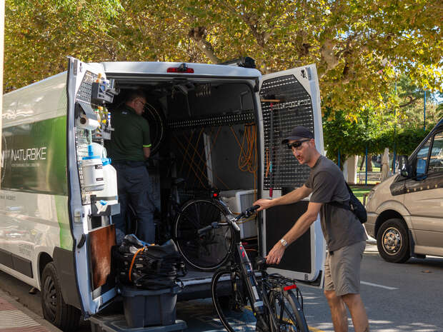 A man holding a bicycle approaches a parked van, where another person adjusts equipment inside. The van is surrounded by leafy trees and another vehicle is nearby.