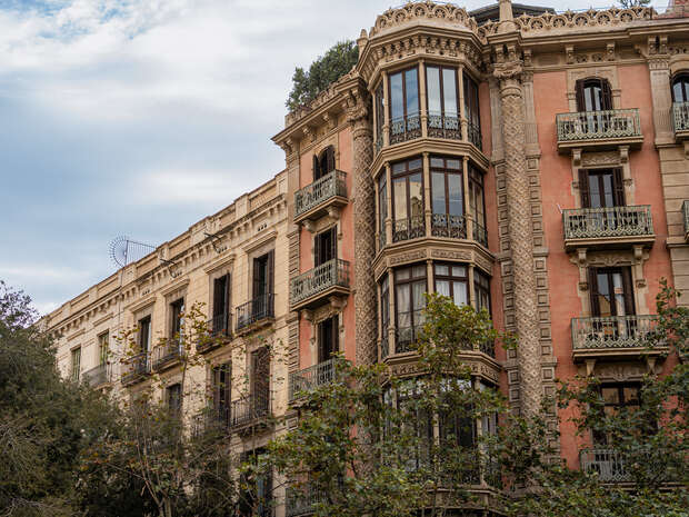 Ornate multi-story building with intricate balconies stands amidst leafy trees; a classic example of European architecture set against a partly cloudy sky.