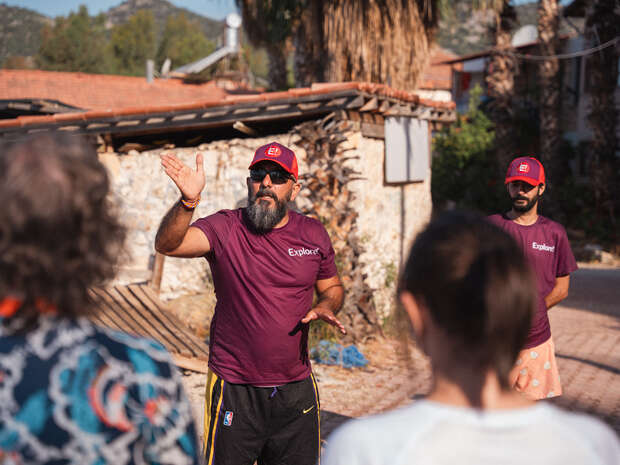 A man wearing a red cap and purple "Explore" shirt gestures with his hand while speaking to a group, outside near a rustic stone building and palm trees.