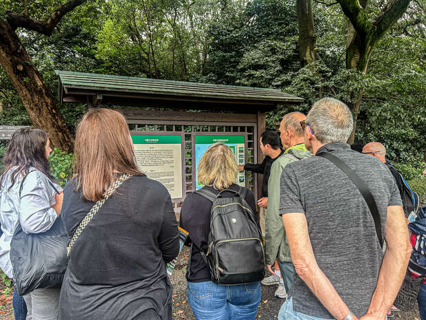 A group of people stand closely, reading information boards under a small shelter surrounded by trees. The boards contain text and images, suggesting educational content about the area.