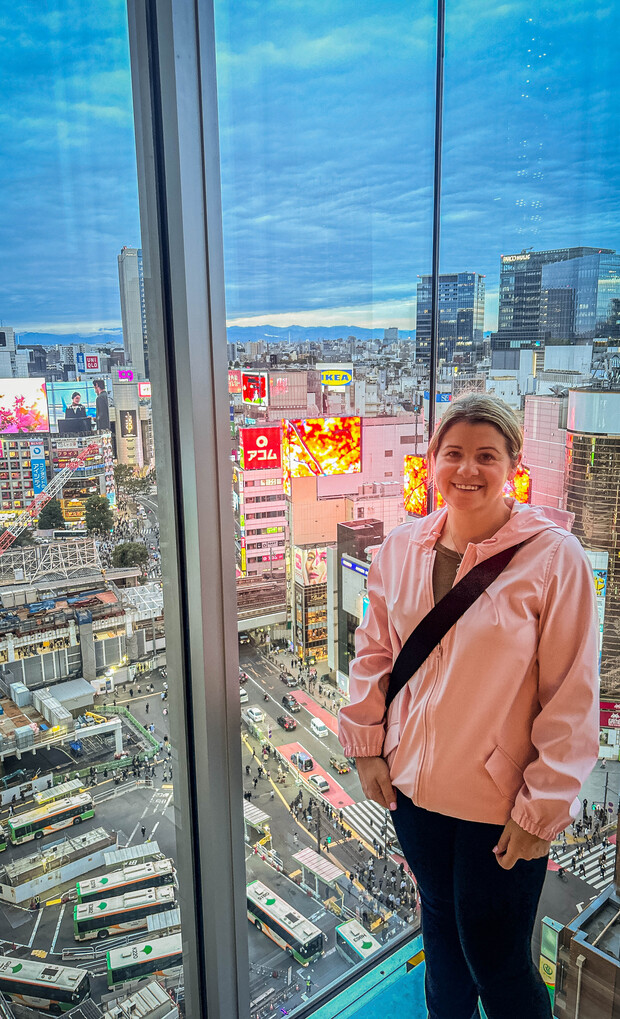 A person in a pink jacket stands by a tall glass window, overlooking a busy city intersection with buses, crowd, and illuminated billboards displaying various advertisements.