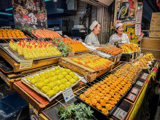 Assorted skewered food is displayed on wooden trays at a bustling market stall. Two people in white uniforms stand behind the counter, smiling. Various signs display prices and product names.