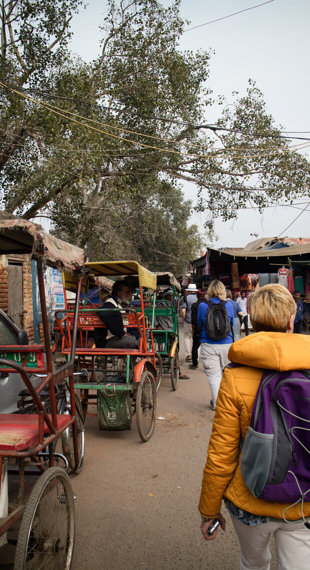 Rickshaws parked on a narrow street, people walking nearby, large leafy trees overhead, with colorful shops lining the road. A person in a yellow jacket and purple backpack walks away.