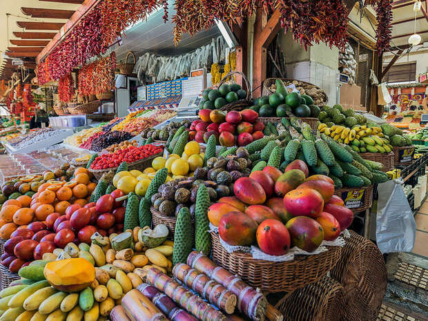 Vibrant fruits, including avocados, mangoes, and bananas, are displayed in baskets at a bustling market. Colorful produce surrounds, with dried peppers hanging above, creating a lively, fresh atmosphere.