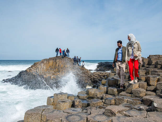 People explore hexagonal basalt columns near the ocean, with waves crashing against the formations. The scene is set under a clear blue sky, where some stand atop higher rocks.