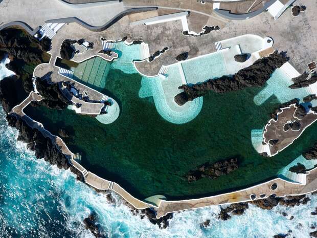 Natural pool complex features turquoise water enclosed by rocky edges, with pathways and platforms for visitors, situated beside the ocean, highlighting a blend of human-made and natural elements.