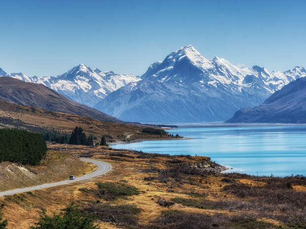 A car drives along a winding road near a tranquil turquoise lake, set against a backdrop of snow-capped mountains under a clear blue sky.