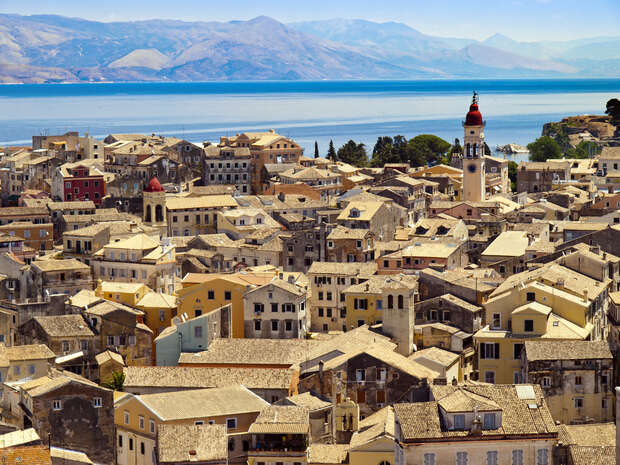 A coastal town with closely packed, colorful houses featuring terracotta roofs rises against a backdrop of blue sea and distant mountains. A prominent clock tower stands amid the buildings.