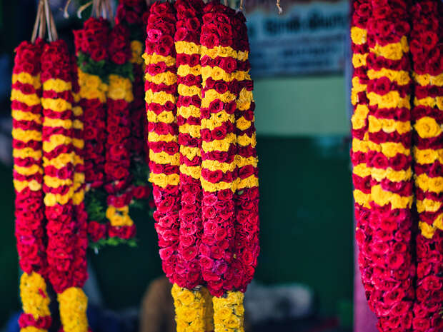Garlands of red and yellow flowers hang in rows. They sway slightly in a market setting, with blurred background elements and a hint of signage in the distance.
